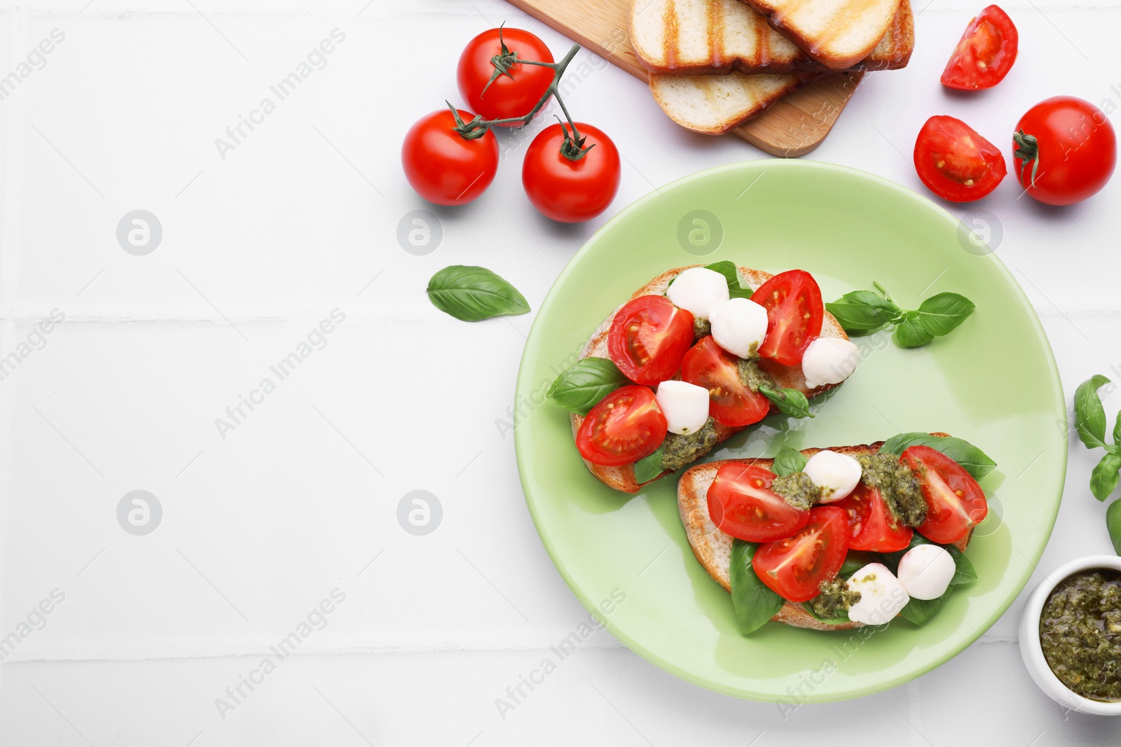 Photo of Delicious Caprese sandwiches with mozzarella, tomatoes, basil and pesto sauce on white tiled table, flat lay. Space for text