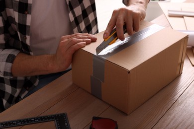 Photo of Man using utility knife to open parcel at wooden table, closeup