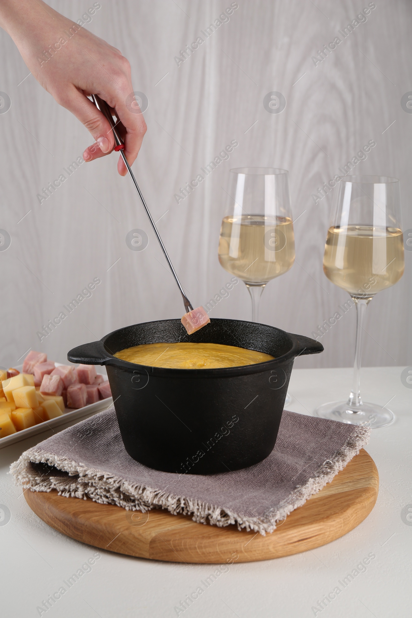 Photo of Dipping piece of ham into fondue pot with tasty melted cheese at white table, closeup