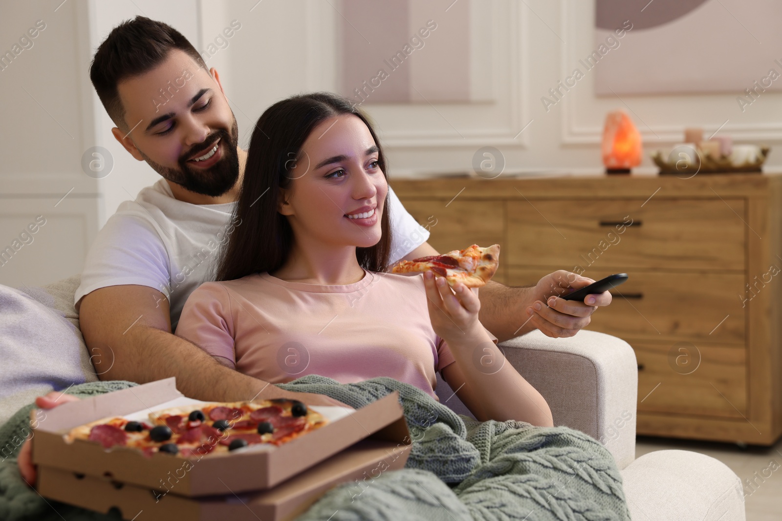 Photo of Happy young couple watching tv and eating pizza on sofa in living room. Space for text