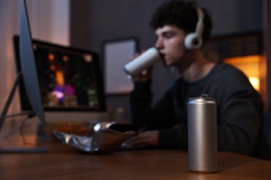 Young man with energy drink playing video game at wooden desk indoors, focus on can
