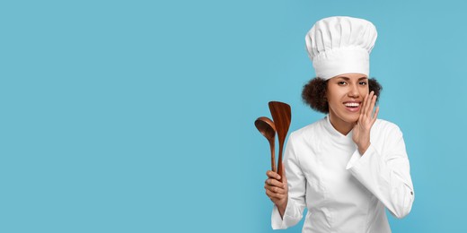 Photo of Happy female chef in uniform holding spoon and spatula on light blue background
