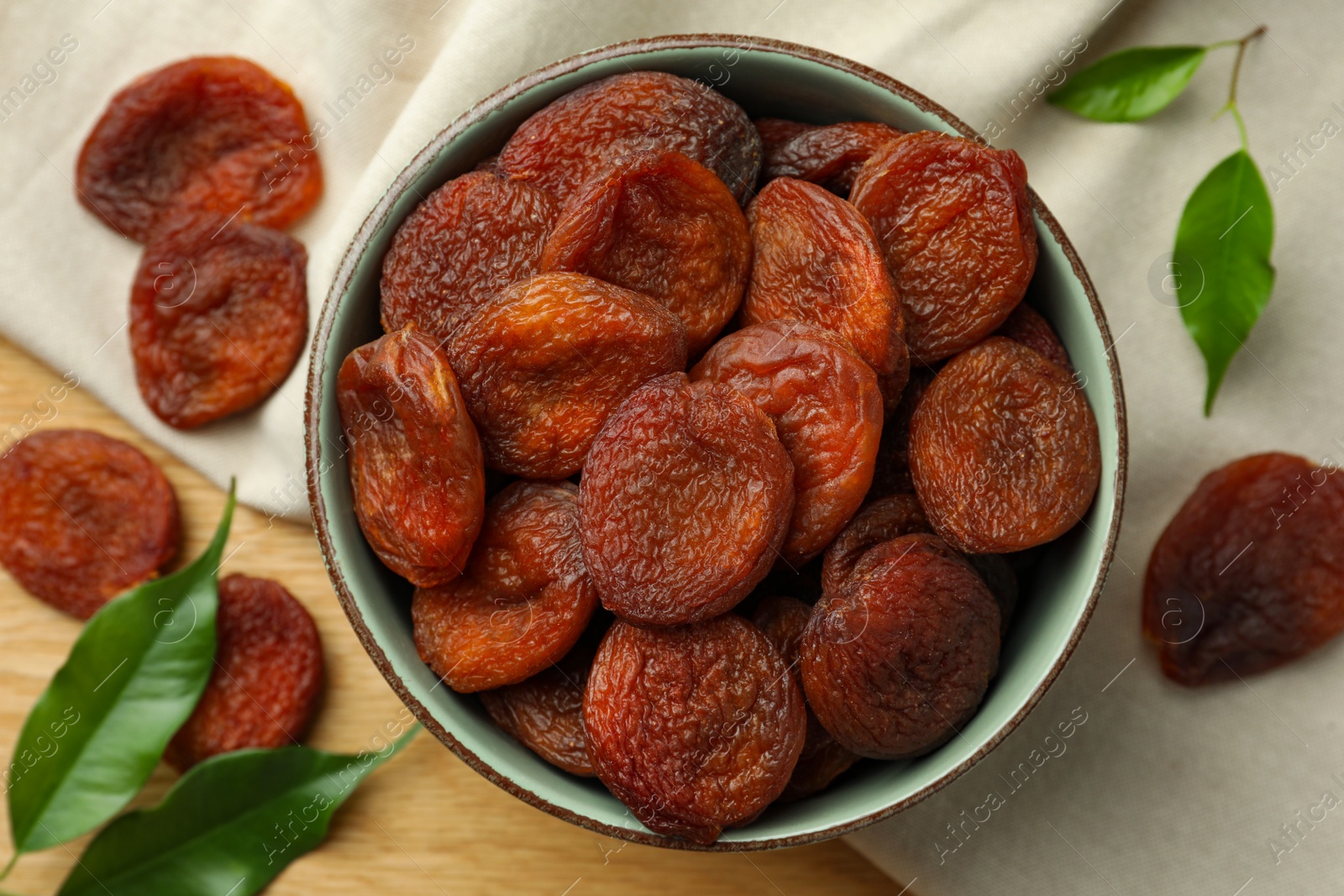 Photo of Tasty apricots with bowl and green leaves on wooden table, flat lay. Dried fruits