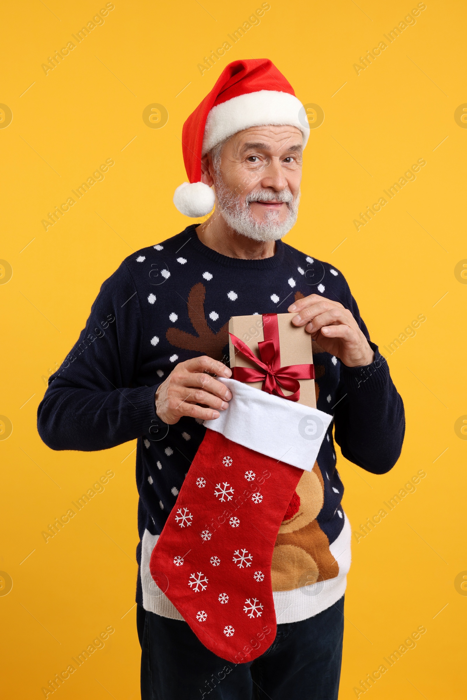 Photo of Senior man in Christmas sweater and Santa hat taking gift from stocking on orange background