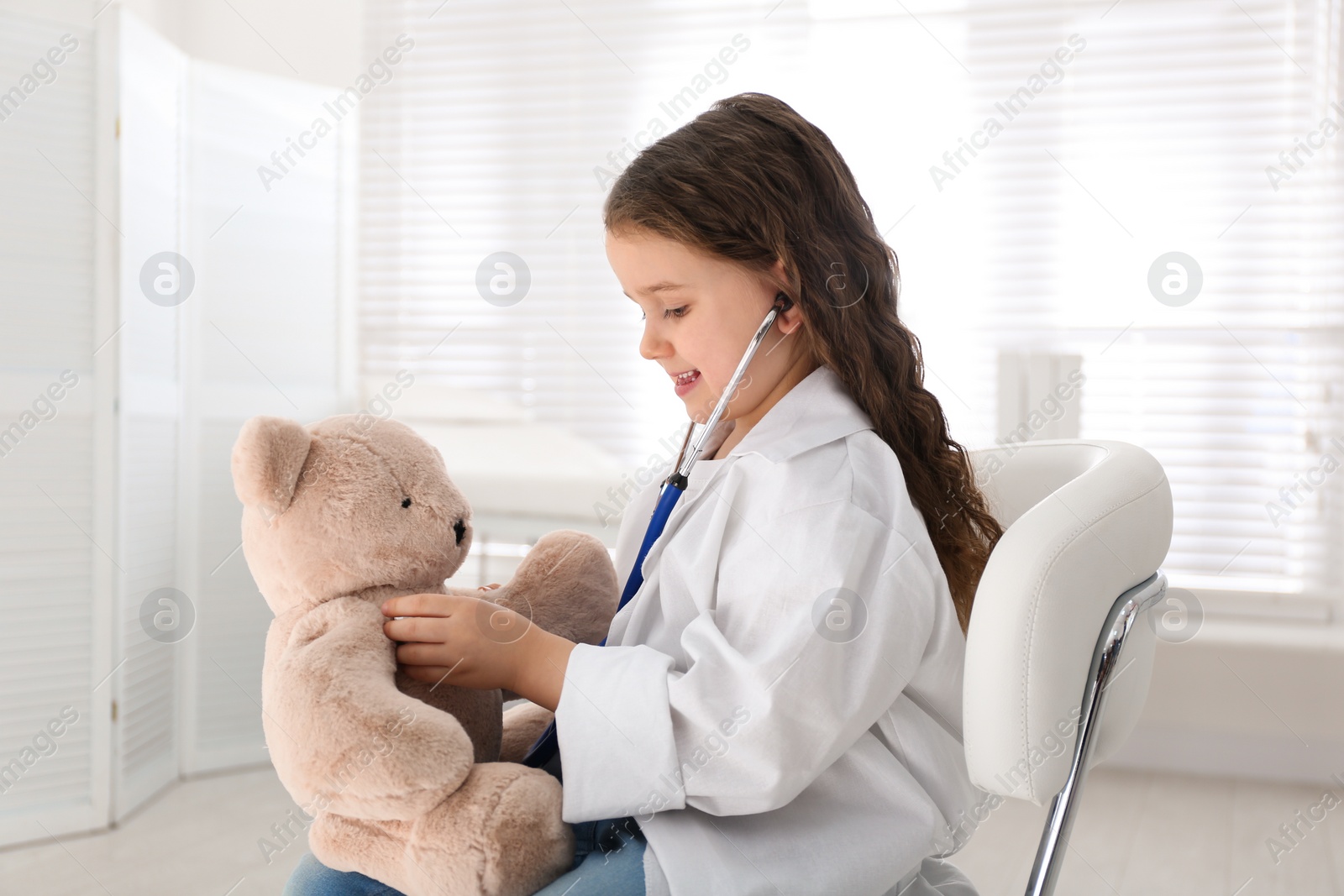 Photo of Cute little girl playing doctor with teddy bear in clinic