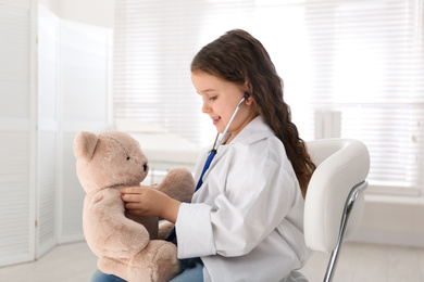 Photo of Cute little girl playing doctor with teddy bear in clinic