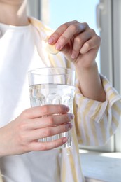 Photo of Woman putting effervescent pill into glass of water indoors, closeup