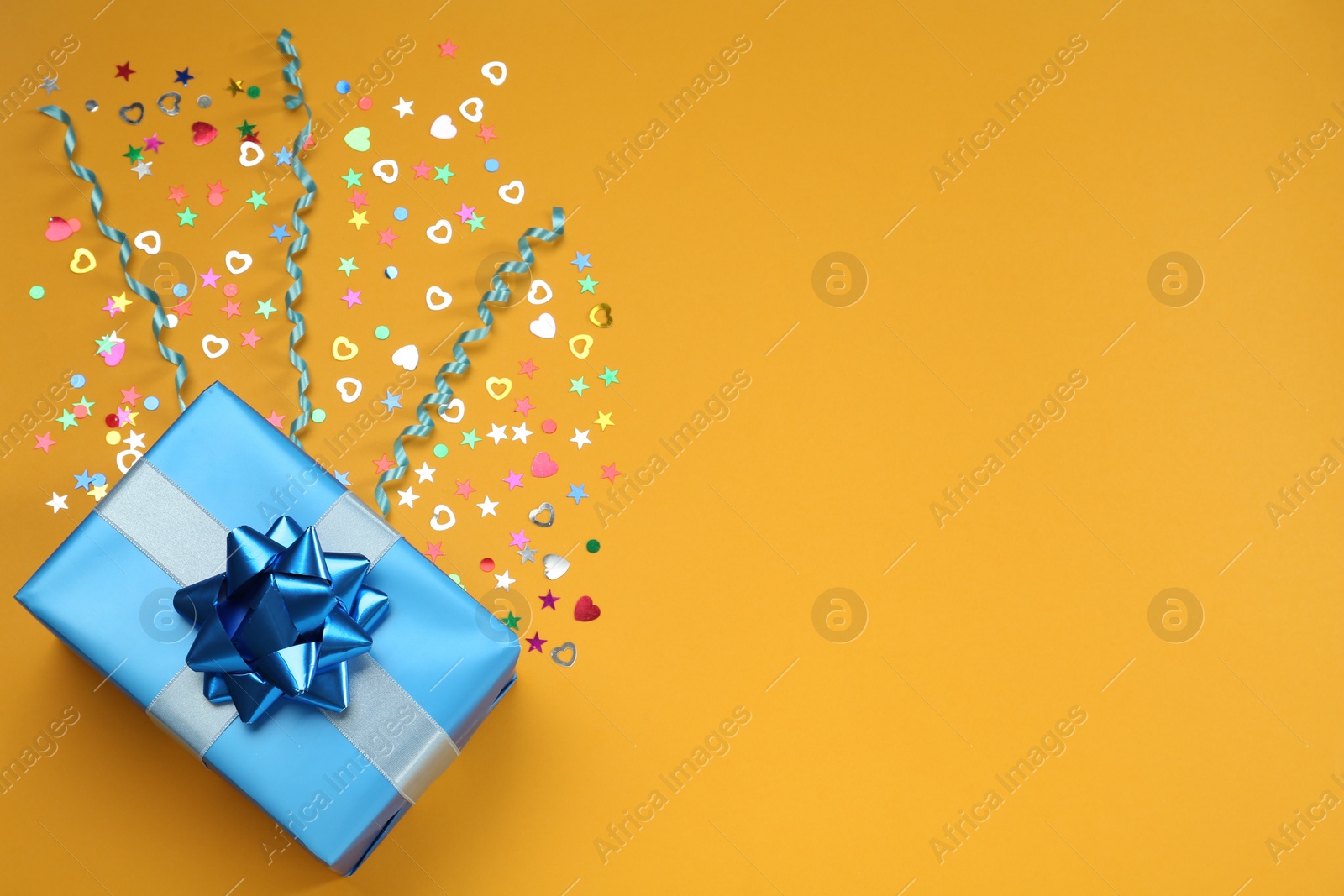 Photo of Light blue gift box with sequins and streamers on orange background, flat lay. Space for text