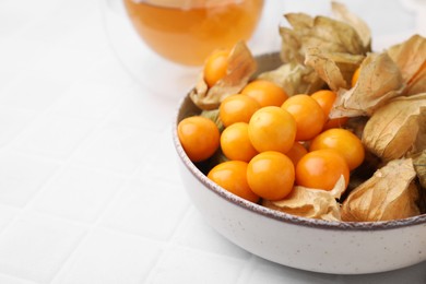 Ripe physalis fruits with calyxes in bowl on white tiled table, closeup. Space for text