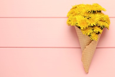 Photo of Beautiful yellow dandelions in waffle cone on pink wooden table, top view. Space for text