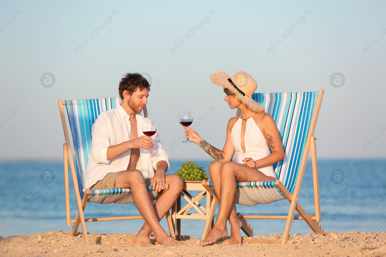 Photo of Young couple with glasses of wine in beach chairs at seacoast