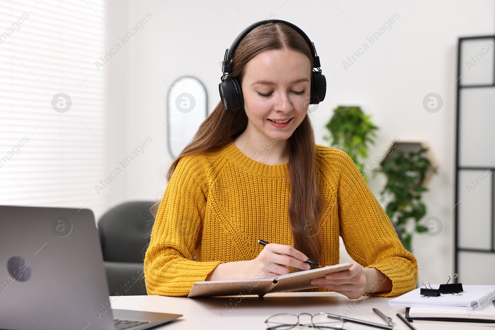 Photo of E-learning. Young woman taking notes during online lesson at white table indoors