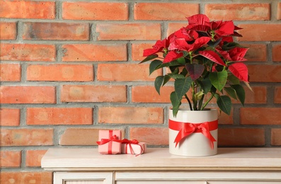 Photo of Beautiful poinsettia (traditional Christmas flower) and gifts on chest of drawers near brick wall. Space for text