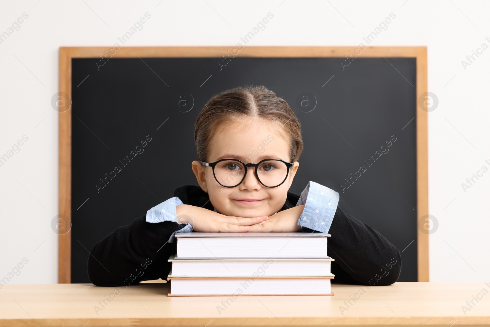 Photo of Happy little school child sitting at desk with books near chalkboard in classroom