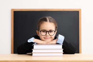 Photo of Happy little school child sitting at desk with books near chalkboard in classroom