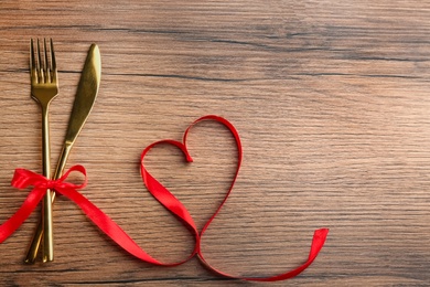 Cutlery set and red ribbon on wooden background, flat lay with space for text. Valentine's Day dinner