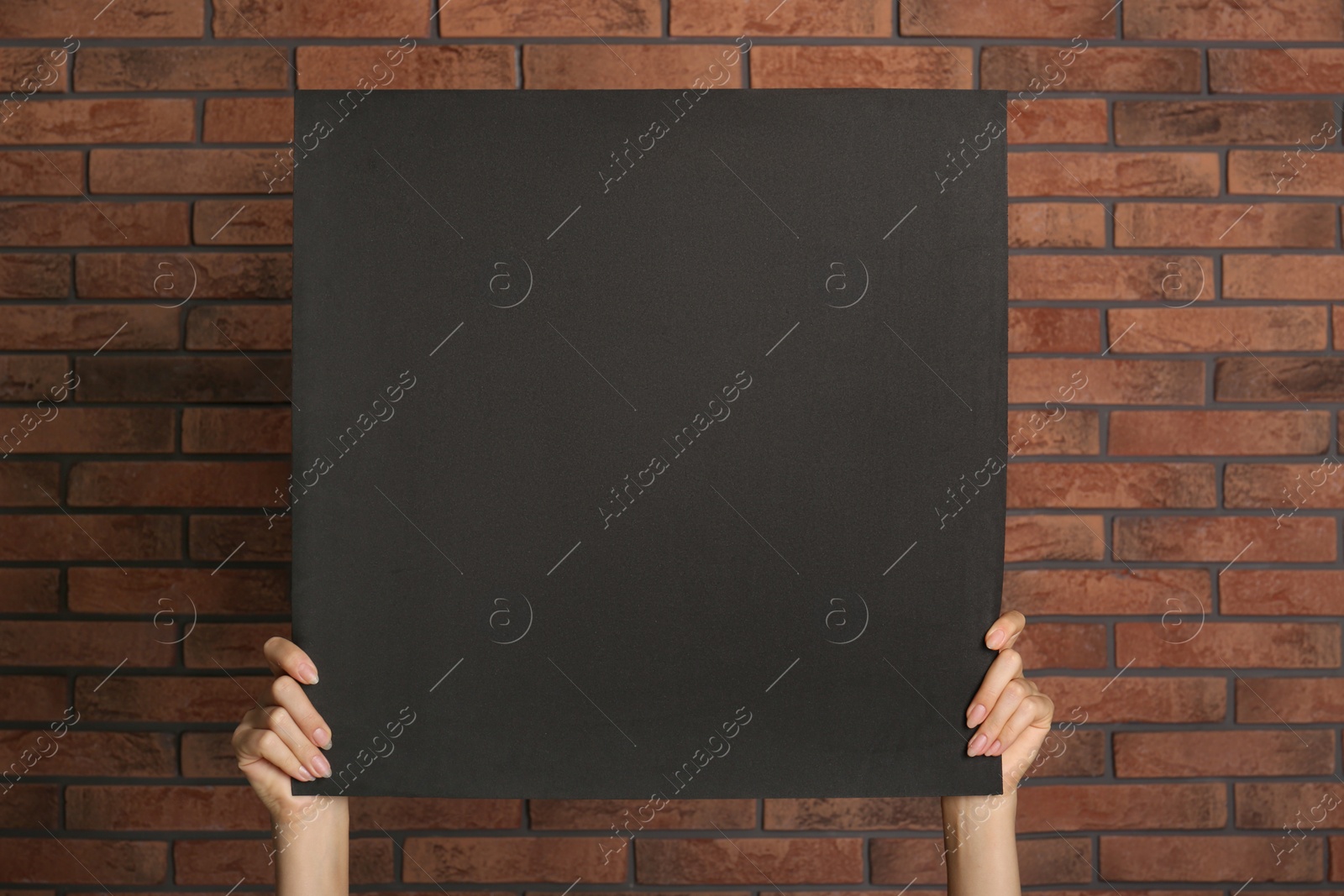 Photo of Woman holding black blank poster near red brick wall, closeup. Mockup for design