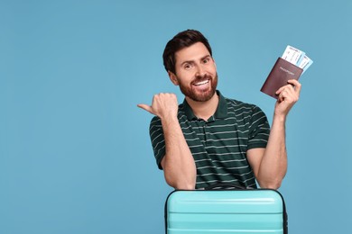 Photo of Smiling man with passport, suitcase and tickets pointing at something on light blue background. Space for text
