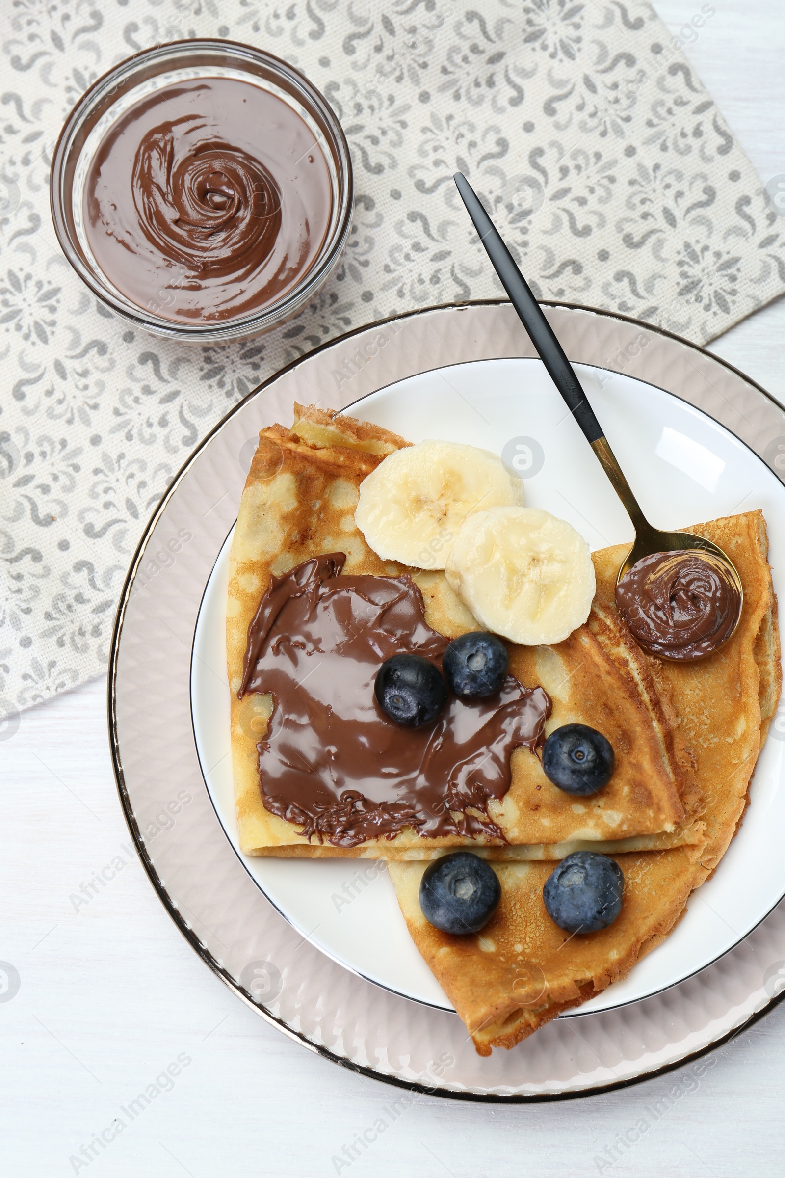 Photo of Tasty crepes with chocolate paste, blueberries and banana served on white wooden table, flat lay