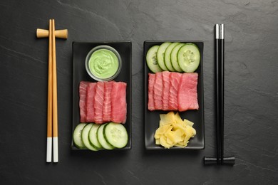 Photo of Tasty sashimi sets (pieces of fresh raw tuna), sliced cucumber, wasabi sauce and ginger slices on black table, flat lay
