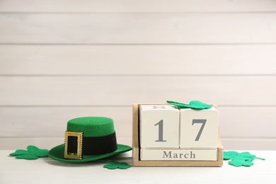 Leprechaun hat, block calendar and decorative clover leaves on white wooden table, space for text. St Patrick's Day celebration