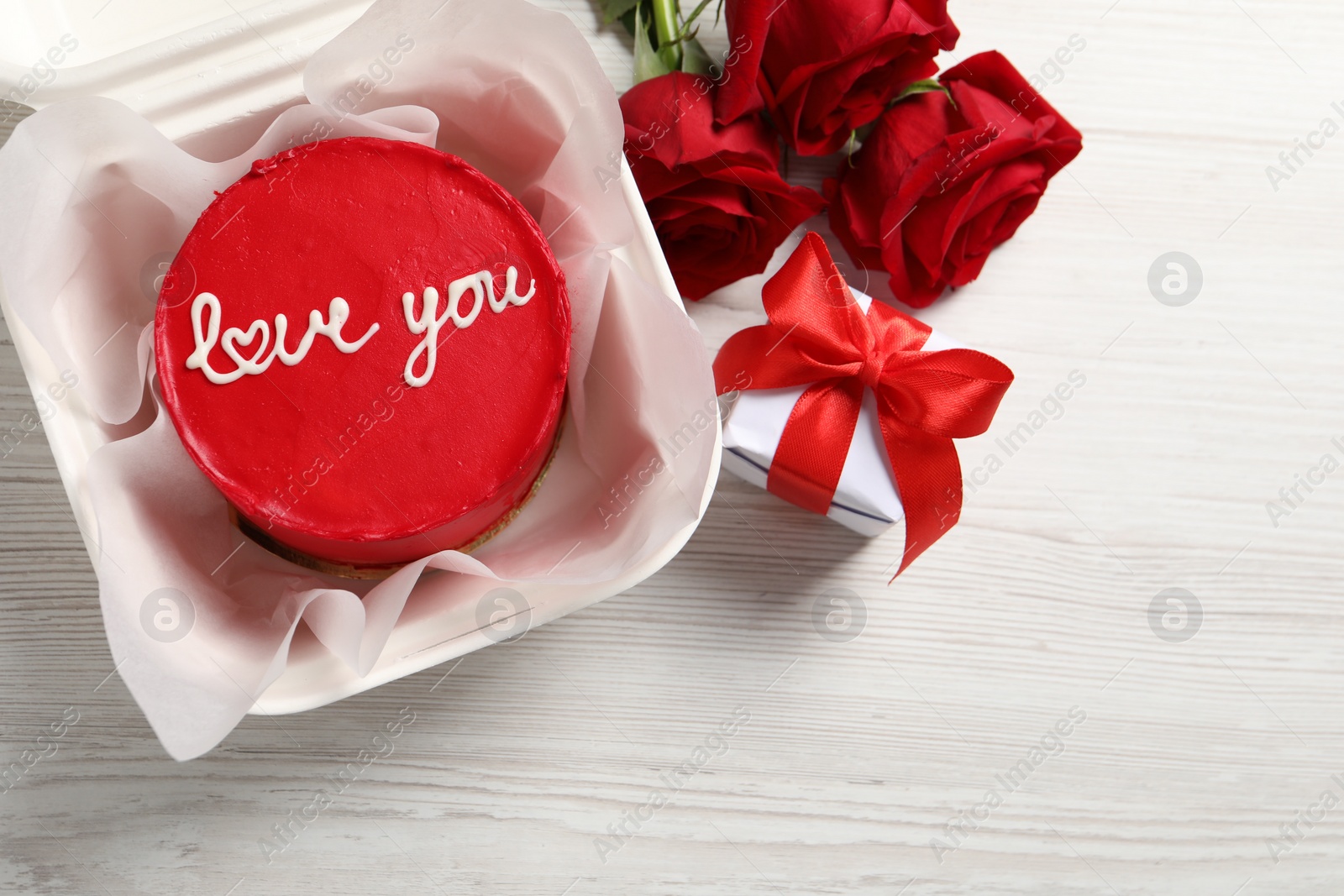 Photo of Bento cake with Love You text in takeaway packaging, roses and gift box on white wooden table, top view. St. Valentine's day surprise