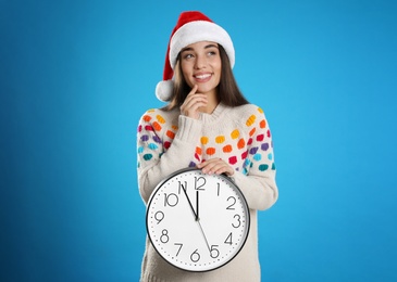 Woman in Santa hat with clock on light blue background. New Year countdown