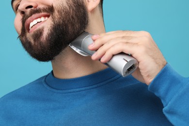 Photo of Handsome young man trimming beard on light blue background, closeup