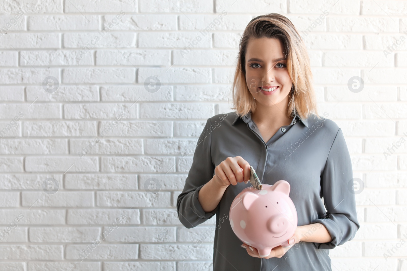 Photo of Beautiful woman putting money into piggy bank near brick wall. Space for text