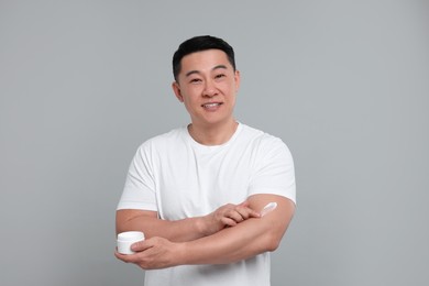 Handsome man applying body cream onto his arm on light grey background