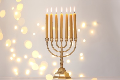Photo of Golden menorah with burning candles against light grey background and blurred festive lights
