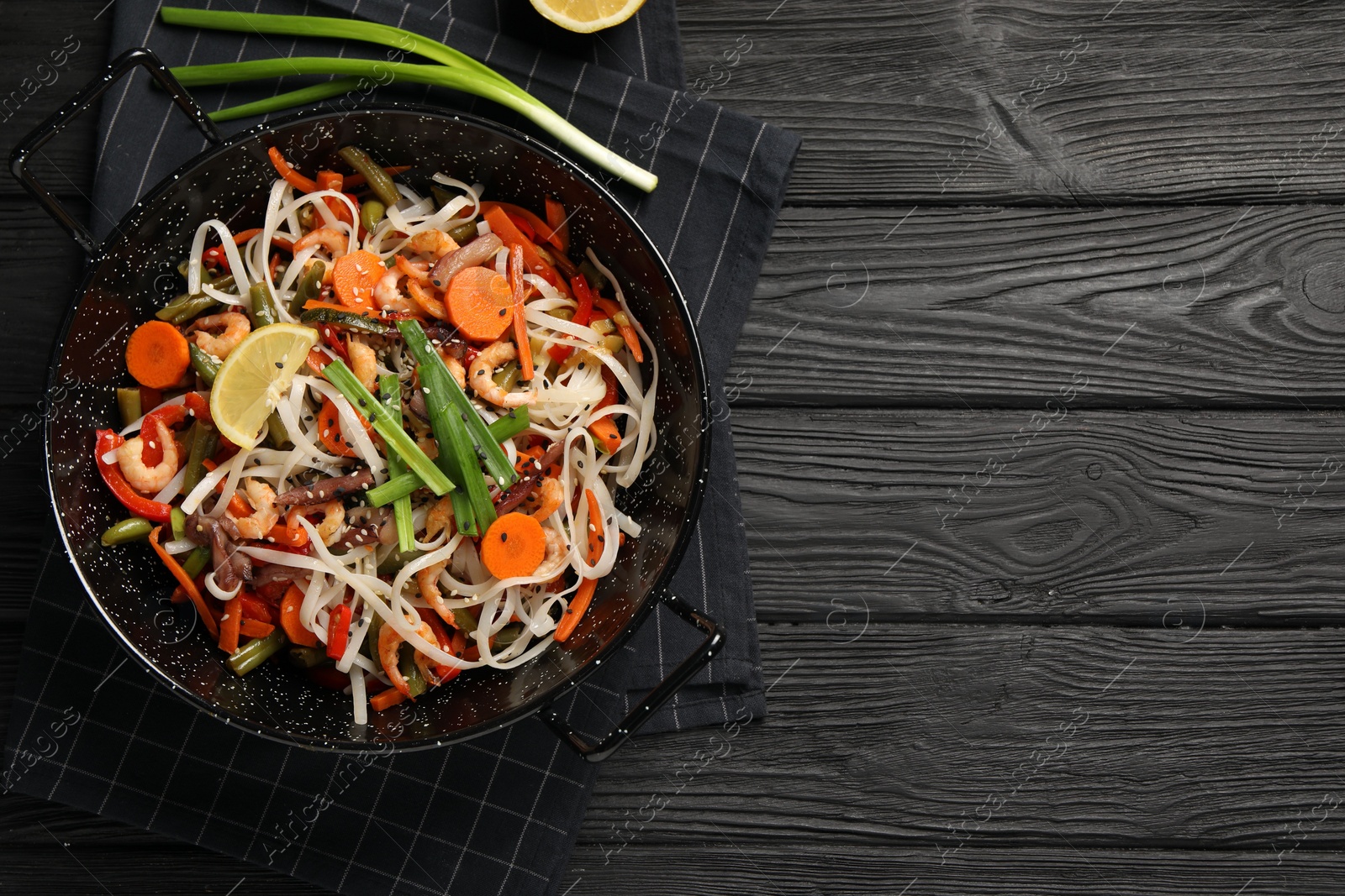 Photo of Shrimp stir fry with noodles and vegetables in wok on black wooden table, top view. Space for text