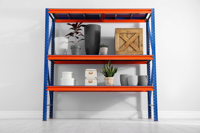 Photo of Metal shelving unit with different household stuff near light wall indoors