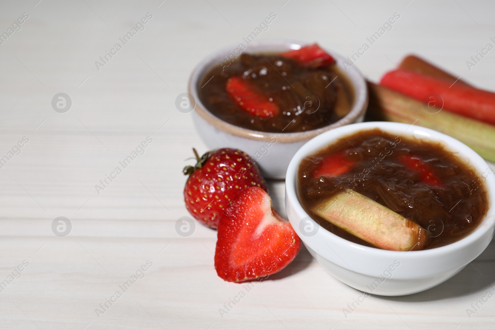 Photo of Tasty rhubarb jam in bowls, stems and strawberries on white wooden table. Space for text