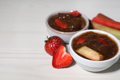 Tasty rhubarb jam in bowls, stems and strawberries on white wooden table. Space for text