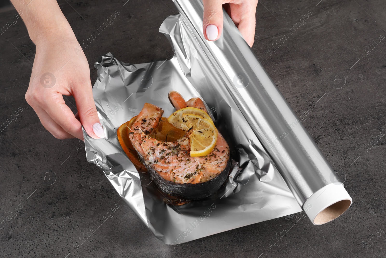 Photo of Woman wrapping tasty salmon with lemon and spices in aluminum foil at grey textured table, closeup