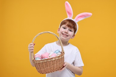 Photo of Easter celebration. Cute little boy with bunny ears and wicker basket full of painted eggs on orange background