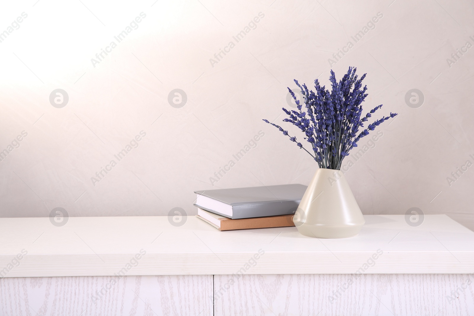 Photo of Bouquet of beautiful preserved lavender flowers and notebooks on white wooden chest of drawers near beige wall, space for text