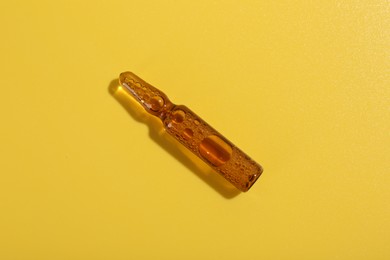 Photo of Glass ampoule with liquid on yellow background, top view