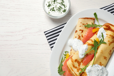 Delicious thin pancakes with salmon and sour cream on white wooden table, flat lay. Space for text
