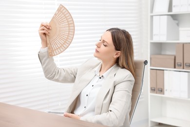 Photo of Businesswoman waving hand fan to cool herself at table in office