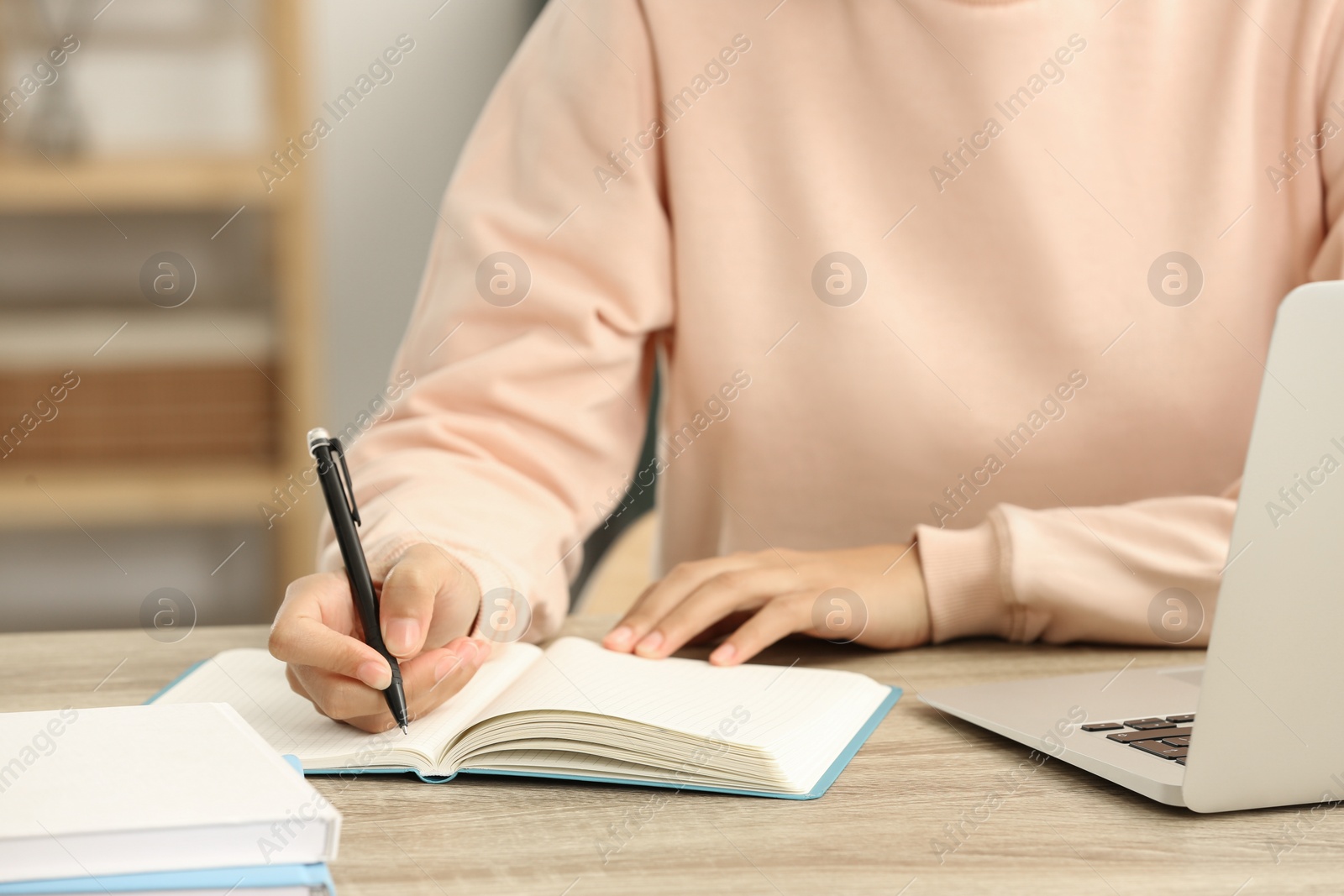 Photo of African American woman writing in notepad near laptop at wooden table indoors, closeup