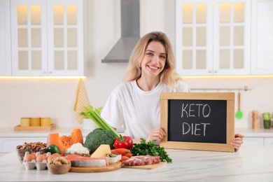 Woman holding blackboard with phrase Keto Diet and different products at white table in kitchen