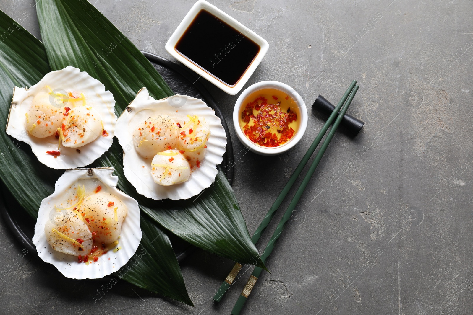 Photo of Raw scallops with spices, lemon zest, shells and sauces on grey textured table, flat lay. Space for text