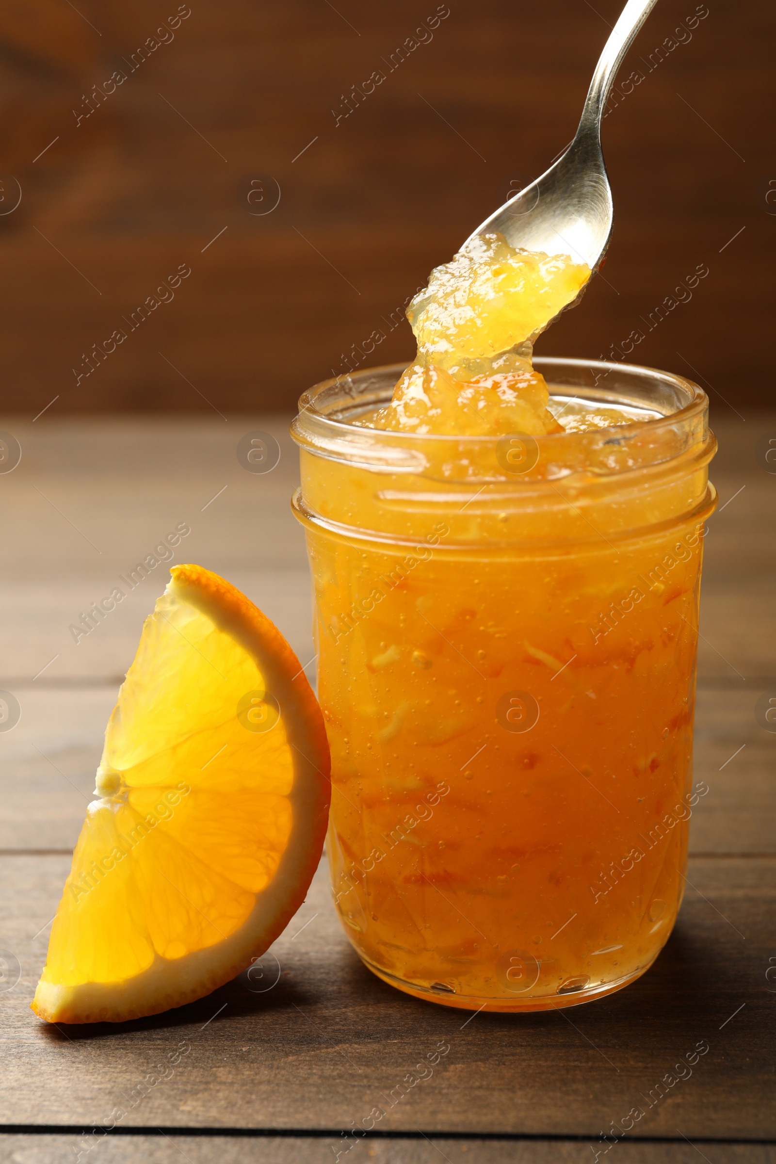 Photo of Spoon with delicious orange marmalade over jar near citrus fruit slice on wooden table