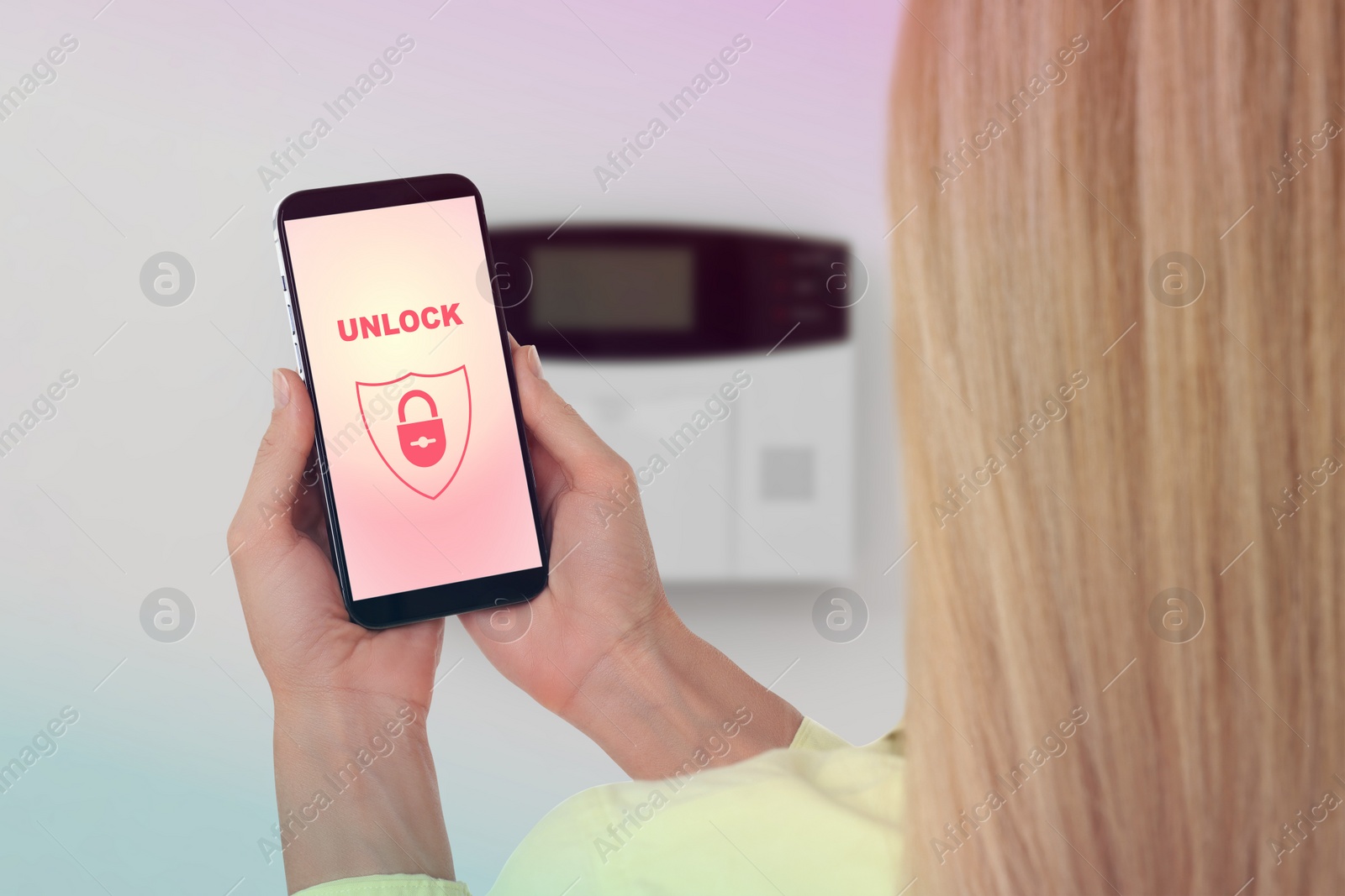 Image of Woman operating home alarm system via mobile phone against white wall with security control panel, closeup. Application interface on device screen