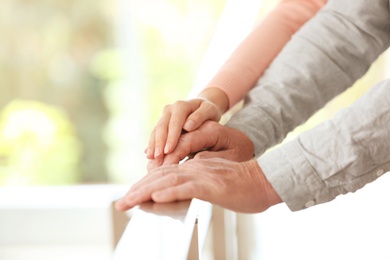 Photo of Young woman holding elderly man hand on railing indoors, closeup. Help service