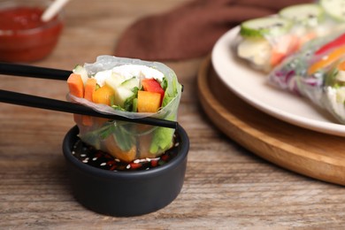 Photo of Dipping delicious roll wrapped in rice paper into soy sauce at wooden table, closeup