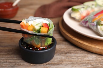 Dipping delicious roll wrapped in rice paper into soy sauce at wooden table, closeup