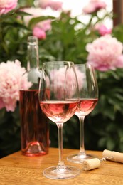 Glasses and bottle with rose wine on wooden table near beautiful peonies